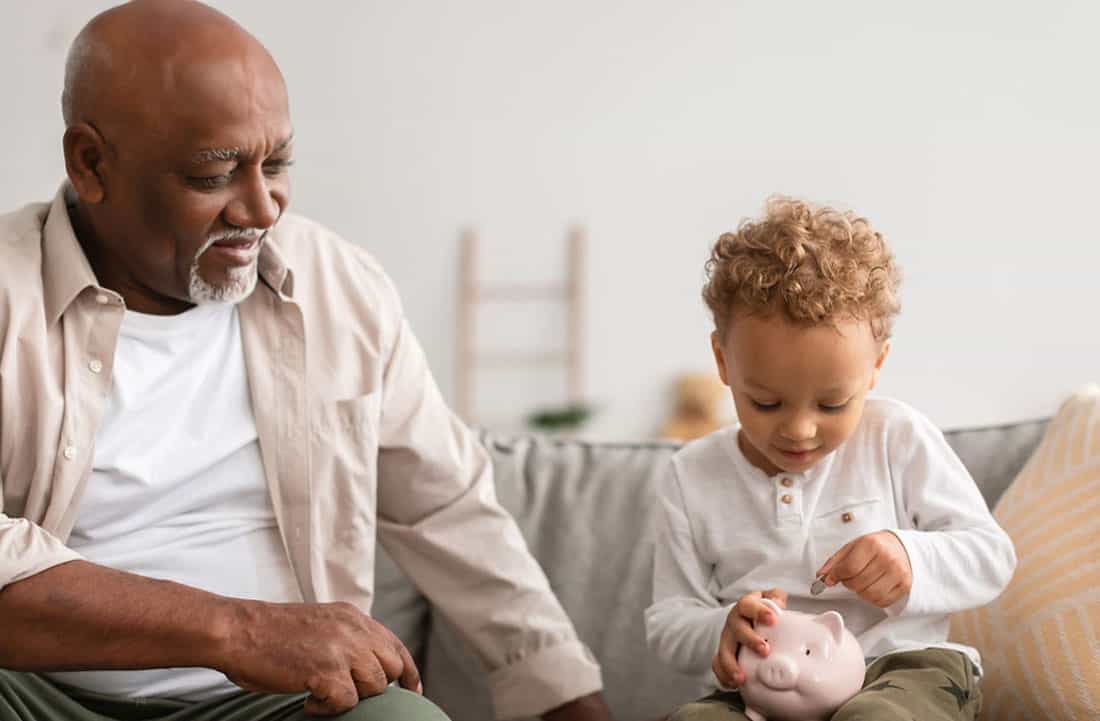 Grandson putting money in a piggy bank while grandfather looks and smiles at him