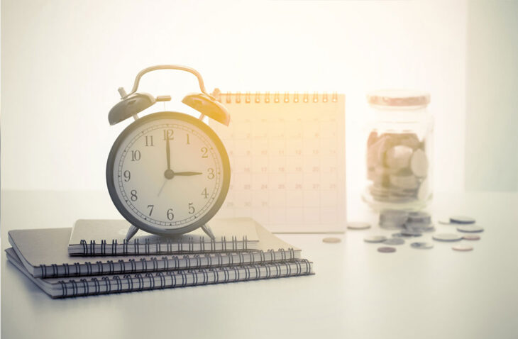 Clock on top of notebooks, calendar, and jar with money in the background