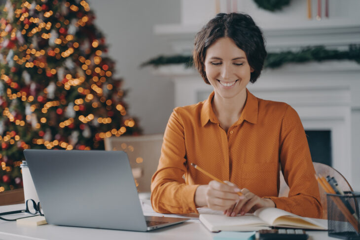 woman smiling at laptop and writing something down on her notebook